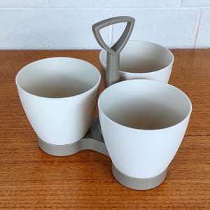 May include: A white plastic condiment caddy with three round compartments. The caddy has a gray handle and base. The caddy is sitting on a wooden surface.