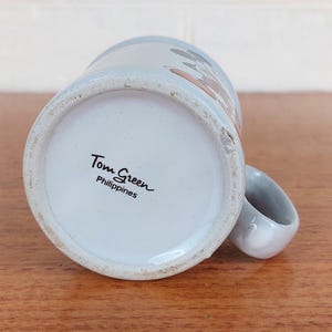 May include: A set of three ceramic mugs with a speckled, matte finish. The mugs are in shades of cream, light gray, and dark gray. Each mug has a simple, minimalist design and a rounded handle.