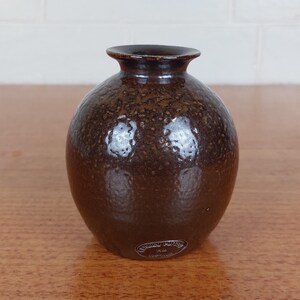 May include: A small, round brown vase with a textured surface. The vase has a narrow neck and a slightly flared rim. The dark brown color has lighter brown speckles throughout. A small label is affixed to the base.