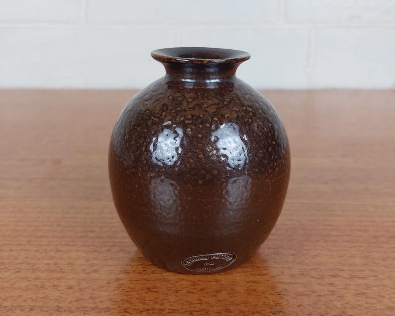 May include: A small, round brown vase with a textured surface. The vase has a narrow neck and a slightly flared rim. The dark brown color has lighter brown speckles throughout. A small label is affixed to the base.