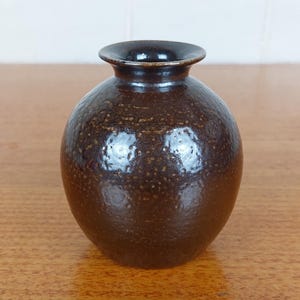May include: A small, round, brown ceramic vase with a dark rim. The vase has a textured surface with small, speckled details. The opening is circular, and the vase sits on a wooden surface. This decorative vase is suitable for home decor.