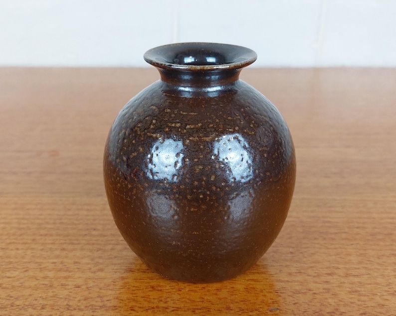 May include: A small, round, brown ceramic vase with a dark rim. The vase has a textured surface with small, speckled details. The opening is circular, and the vase sits on a wooden surface. This decorative vase is suitable for home decor.
