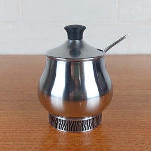 May include: A stainless steel sugar bowl with a bulbous body, a removable lid featuring a black knob, and a small metal spoon. The base of the bowl is decorated with a dark, vertical, abstract line pattern that encircles the bottom perimeter.