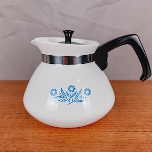 Puede incluir: Tetera de vidrio blanco con asa y tapa negras. La tetera presenta una banda plateada y un diseño floral azul. El asa es curva y la tapa tiene un pequeño pomo negro.