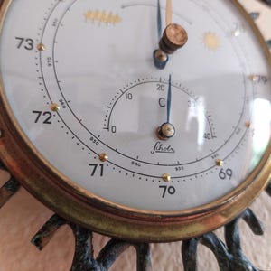 May include: Close-up of a vintage barometer with a white face, gold-colored trim, and a decorative bronze frame. The barometer displays temperature in Celsius and pressure readings. The word "Scholz" is printed on the face.