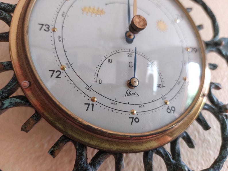May include: Close-up of a vintage barometer with a white face, gold-colored trim, and a decorative bronze frame. The barometer displays temperature in Celsius and pressure readings. The word "Scholz" is printed on the face.