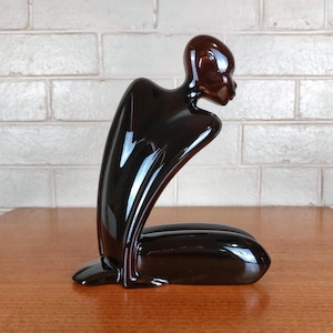 May include: A glossy, dark brown ceramic sculpture of a stylised figure in a kneeling, forward-leaning posture. The smooth, reflective surface highlights the abstract, elongated form of the body, which is depicted without facial features.