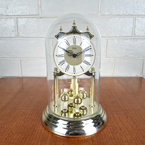 May include: A gold-tone anniversary clock encased in a clear glass dome. The clock features a white face with black Roman numerals, black hands, and a central clover-shaped detail. It stands on a circular gold base with four rotating pendulums.