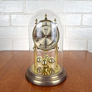 May include: A gold-toned quartz anniversary clock with a circular face, black Roman numerals, and black hands, enclosed within a clear glass dome. The clock features a rotating pendulum with four gold spheres and sits on a circular base.