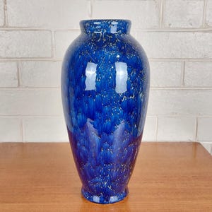 Puede incluir: Un jarrón azul alto con un acabado brillante. El jarrón tiene un patrón moteado de blanco y azul, con una base acampanada y un cuello estrecho. El jarrón está sobre una mesa de madera.