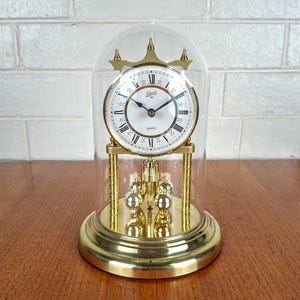 May include: A gold-tone quartz anniversary clock with a white face, black Roman numerals, and black hands, enclosed in a clear glass dome. The clock features a rotating pendulum with four gold-tone spheres and stands on a circular gold base.