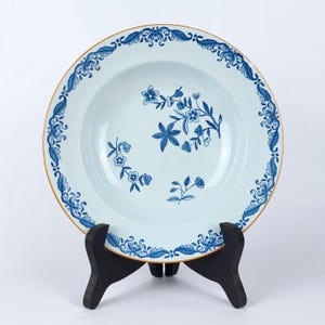 Puede incluir: Un tazón de sopa de cerámica blanca con un diseño floral azul y dorado. El tazón tiene un borde festoneado y está sobre un soporte negro.