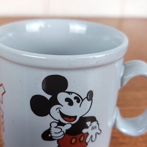 May include: A light blue ceramic mug featuring a smiling Mickey Mouse graphic. The mug has a rounded handle and a speckled finish. The iconic Disney character is depicted in black, red, and white.