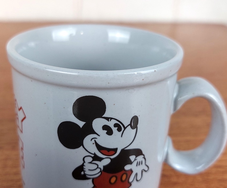 May include: A light blue ceramic mug featuring a smiling Mickey Mouse graphic. The mug has a rounded handle and a speckled finish. The iconic Disney character is depicted in black, red, and white.