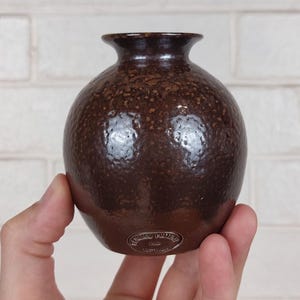 May include: A small, round, brown ceramic vase with a textured surface. The vase has a narrow neck and a slightly flared rim. The bottom of the vase has a small, circular stamp with text. The vase is held in a hand.