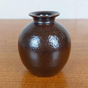 May include: A small, round, brown ceramic vase with a textured surface. The vase has a narrow neck and a dark brown interior. The exterior has a speckled appearance, and the vase is sitting on a wooden surface.