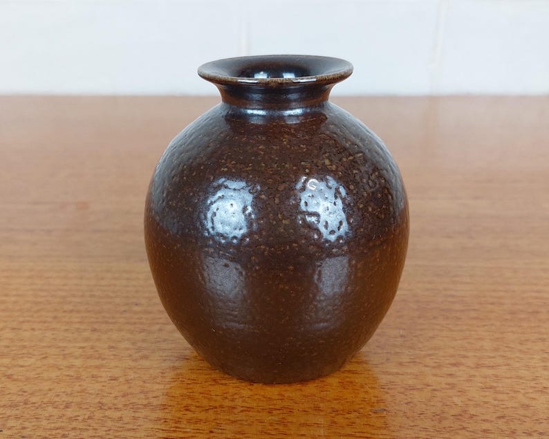 May include: A small, round, brown ceramic vase with a textured surface. The vase has a narrow neck and a dark brown interior. The exterior has a speckled appearance, and the vase is sitting on a wooden surface.