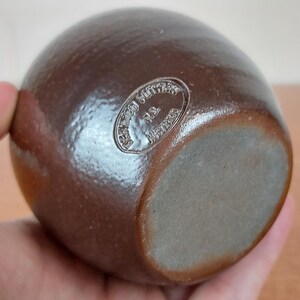May include: A brown, egg-shaped ceramic vase with a matte finish. The vase has a rounded body and a flat base. The bottom of the vase has a circular stamp that reads "Bennington Pottery P.L. Vermont".