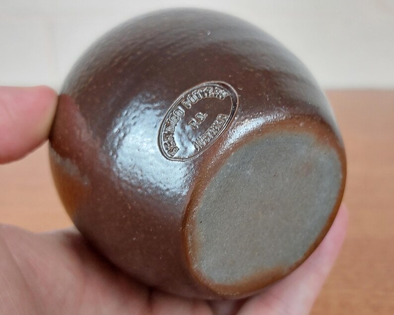 May include: A brown, egg-shaped ceramic vase with a matte finish. The vase has a rounded body and a flat base. The bottom of the vase has a circular stamp that reads "Bennington Pottery P.L. Vermont".
