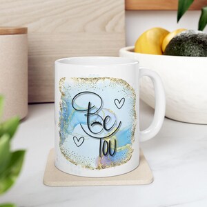 Puede incluir: Taza de cerámica blanca con la frase "Be You" en escritura negra, acentuada con un diseño de acuarela azul y dorada y tres pequeños corazones. La taza tiene un asa estándar y está sobre un posavasos beige.