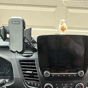 May include: Two black and gray adjustable smartphone mounts attached to the air vents of a vehicle dashboard, flanking a central infotainment screen. A small, fuzzy, light brown figurine sits on top of the screen, and a white USB cable hangs down.