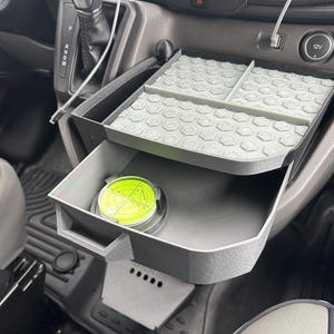 May include: A dark gray plastic vehicle dashboard storage organizer with two tiers. The top tier features a light gray hexagonal patterned mat divided into three sections. The bottom tier is a pull-out drawer containing a circular yellow spirit level.