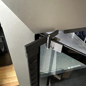 以下が含まれることがあります： グレーの金属製のドアに取り付けられた、黒色の3Dプリントされたヒンジのクローズアップ。ドアはテクスチャ加工された表面と黒いトリムを持っています。ヒンジは小さなネジでドアに取り付けられています。