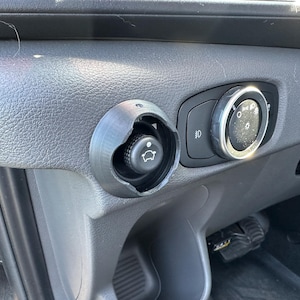 May include: Close-up of a vehicle's dashboard featuring a black, circular control knob with a light symbol and a smaller, round control with a car symbol. The dashboard is gray and textured, with additional light controls.