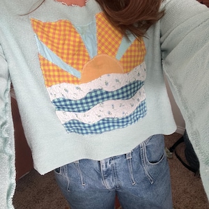 K&ouml;nnte beinhalten: Ein helles mintgr&uuml;nes Langarm-Sweatshirt mit offenem Halsausschnitt und Saum. In der Mitte befindet sich eine Stoffapplikation einer aufgehenden Sonne &uuml;ber Wellen, gefertigt aus gelb-orangefarbenem Vichy-Karo, Hellblau und blau-wei&szlig;em Blumenstoff.