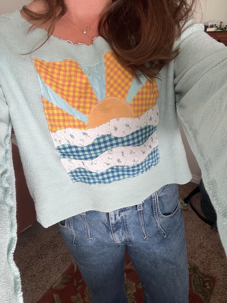 K&ouml;nnte beinhalten: Ein helles mintgr&uuml;nes Langarm-Sweatshirt mit offenem Halsausschnitt und Saum. In der Mitte befindet sich eine Stoffapplikation einer aufgehenden Sonne &uuml;ber Wellen, gefertigt aus gelb-orangefarbenem Vichy-Karo, Hellblau und blau-wei&szlig;em Blumenstoff.