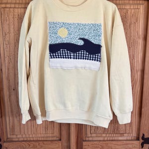May include: A pale yellow sweatshirt with a crew neck and long sleeves. The front features a patchwork design of a blue and white floral pattern, a navy blue wave, a yellow circle, and a black and white checkered pattern.