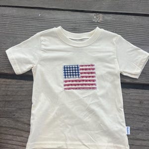May include: A cream-coloured short-sleeve t-shirt featuring a rectangular patch on the chest. The patch displays a stylised American flag design with a blue and white gingham canton and red and white striped sections, all finished with visible stitching.