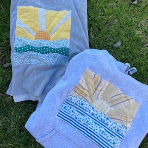 K&ouml;nnte beinhalten: Ein hellgraues Sweatshirt mit Rundhalsausschnitt und ein hellgrauer Kapuzenpullover, jeweils mit einem rechteckigen Stoffaufn&auml;her. Die Aufn&auml;her zeigen eine aufgehende Sonne &uuml;ber einer Landschaft, zusammengesetzt aus gemusterten Stoffresten in Gelb, Blau und Gr&uuml;n.