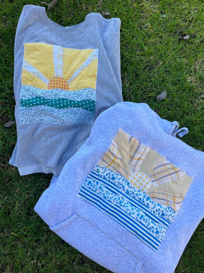 K&ouml;nnte beinhalten: Ein hellgraues Sweatshirt mit Rundhalsausschnitt und ein hellgrauer Kapuzenpullover, jeweils mit einem rechteckigen Stoffaufn&auml;her. Die Aufn&auml;her zeigen eine aufgehende Sonne &uuml;ber einer Landschaft, zusammengesetzt aus gemusterten Stoffresten in Gelb, Blau und Gr&uuml;n.