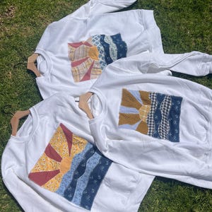 K&ouml;nnte beinhalten: Drei wei&szlig;e Sweatshirts mit Rundhalsausschnitt, die jeweils eine rechteckige Stoffapplikation auf der Brust aufweisen. Die Applikation zeigt eine aufgehende Sonne &uuml;ber wellenf&ouml;rmigen Landschaftsmustern aus verschiedenen gemusterten Stoffst&uuml;cken in Blau, Gelb und Rot.