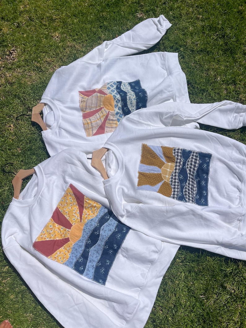 K&ouml;nnte beinhalten: Drei wei&szlig;e Sweatshirts mit Rundhalsausschnitt, die jeweils eine rechteckige Stoffapplikation auf der Brust aufweisen. Die Applikation zeigt eine aufgehende Sonne &uuml;ber wellenf&ouml;rmigen Landschaftsmustern aus verschiedenen gemusterten Stoffst&uuml;cken in Blau, Gelb und Rot.
