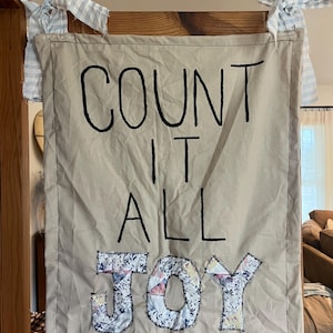 May include: A rectangular beige fabric wall hanging featuring the black embroidered text COUNT IT ALL, with the word JOY appliqued in a multi-colored floral fabric. The banner is suspended by two light blue and white gingham fabric ties.