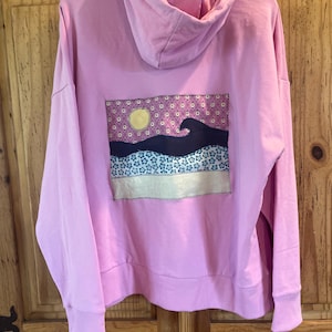 May include: A light pink hoodie with a hood and long sleeves. The back of the hoodie features a rectangular patch with a landscape design. The design includes a yellow sun, a dark blue wave, and a starry sky.