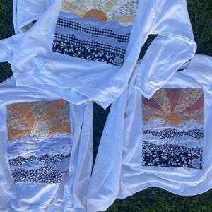 May include: Three white sweatshirts with a patchwork design of a sunrise over the ocean. The sun is a mix of orange, yellow, and brown fabrics. The ocean is represented by blue and navy fabrics. The sweatshirts are on a green grassy surface.