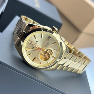 May include: A gold-toned Bulova automatic watch with a matching link bracelet. The watch face features a skeleton design, revealing the inner workings. The watch has a gold dial with hour markers and hands. The watch is displayed on a dark gray surface.