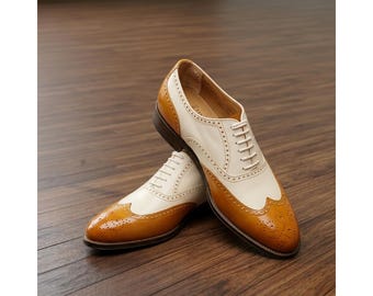 May include: A pair of two-tone wingtip dress shoes. The shoes are cream-colored with tan accents on the toe and heel. The shoes have a classic lace-up design and are made of leather. The shoes are displayed on a wooden surface.