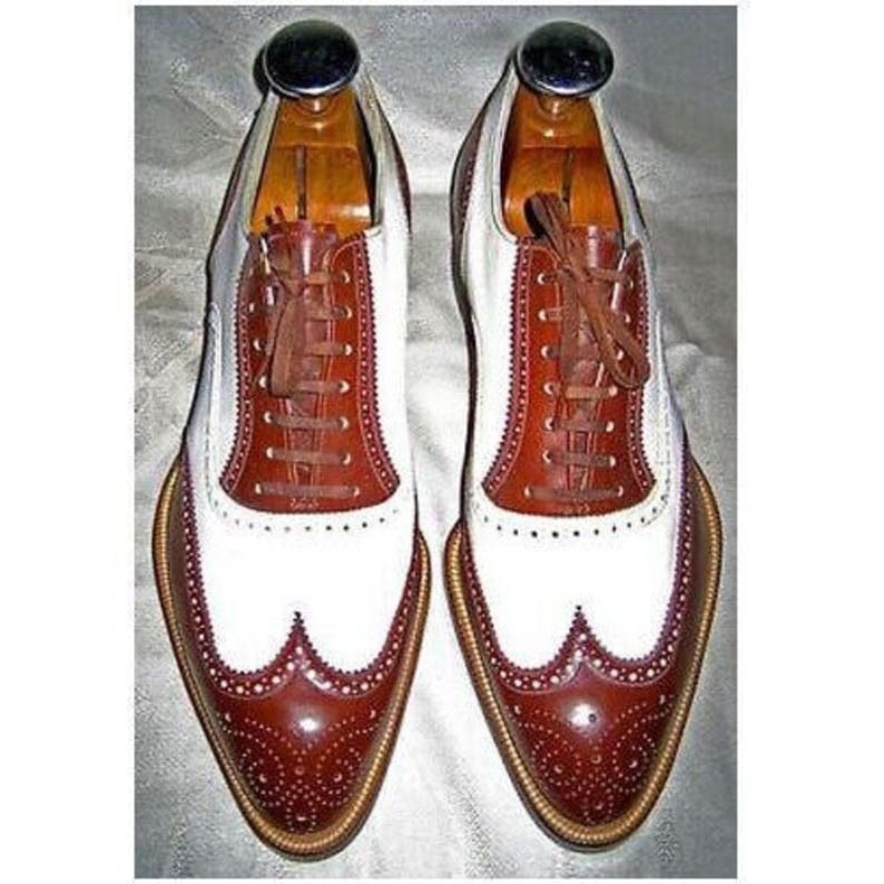 May include: A pair of two-tone leather dress shoes with brown and white panels. The shoes feature brown laces, decorative perforations, and a classic wingtip design. Wooden shoe trees are inserted inside.
