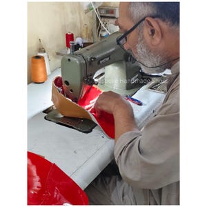 May include: A person using a Brother sewing machine to stitch together the red and tan leather components of a shoe. The text "Bespoke Handmade Shoes" is visible on the machine.