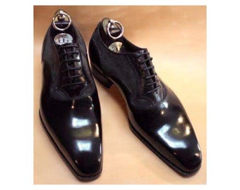 May include: A pair of black leather oxford shoes featuring a high-shine polished finish on the toe and heel, contrasted with a matte textured panel on the side. Each shoe includes black laces and a silver-toned metal shoe tree with a ring handle.
