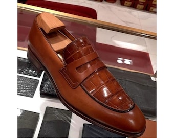 May include: A tan leather loafer featuring a smooth leather heel and side panel, contrasted with a textured crocodile-embossed leather vamp and strap. A wooden shoe tree is inserted into the opening of the shoe, which is displayed on a glass shelf.