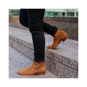 May include: A pair of tan suede ankle boots with a low heel. The boots have a rounded toe and a folded design around the ankle. The boots are worn with black denim jeans. The boots are made of suede.