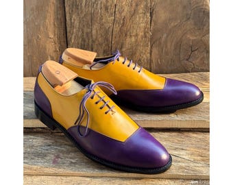May include: A pair of two-tone leather dress shoes. The shoes feature a yellow vamp and quarter, with a purple toe cap and heel. The shoes have purple laces and are set on a wooden surface. Shoe trees are inserted in the shoes.