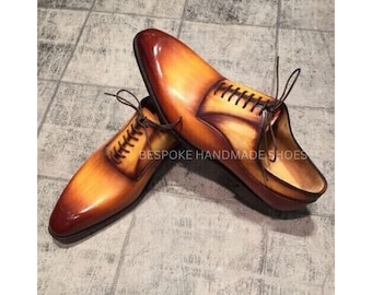 May include: A pair of handcrafted, two-tone leather dress shoes. The shoes feature a gradient color scheme, transitioning from a deep burgundy at the toe to a light tan in the center. The text "BESPOKE HANDMADE SHOES" is visible.