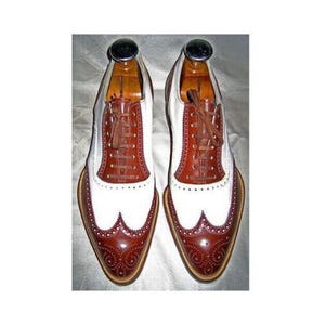 May include: A pair of two-tone leather dress shoes with brown and white panels. The shoes feature a wingtip design with decorative perforations and brown laces. The shoes are on wooden shoe trees.