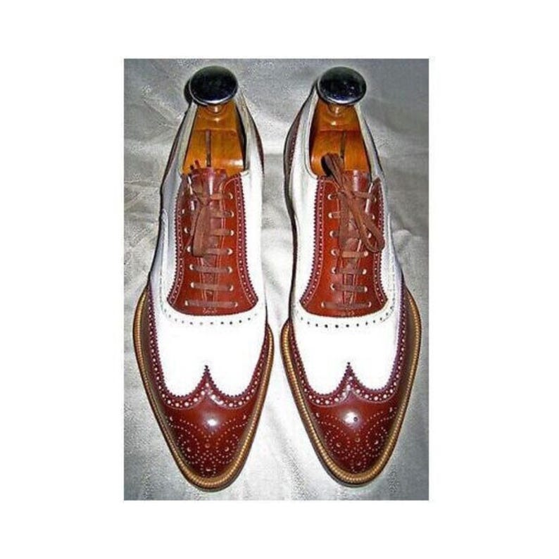 May include: A pair of two-tone leather dress shoes with brown and white panels. The shoes feature a wingtip design with decorative perforations and brown laces. The shoes are on wooden shoe trees.